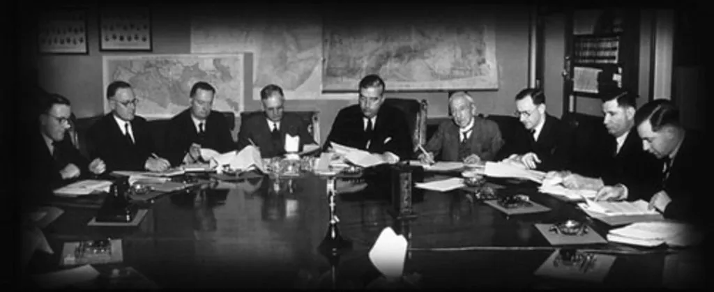 Prime Minister Robert Menzies sits at a table, with four men sitting on each side, all looking at papers, large maps pinned to the wall in the background. Prime Minister Robert Menzies sits at a table, with four men sitting on each side, all looking at papers, large maps pinned to the wall in the background.