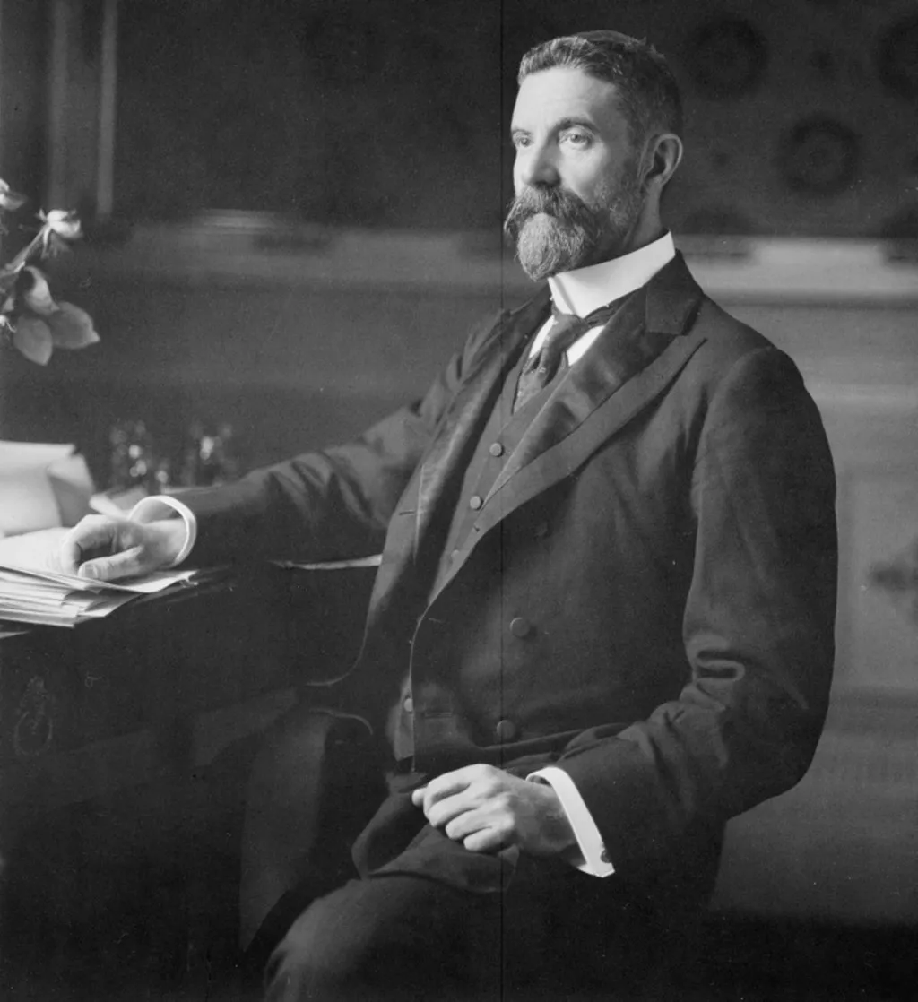 Alfred Deakin, wearing a three-piece suit and seated at a desk covered in paper documents. Alfred Deakin, wearing a three-piece suit and seated at a desk covered in paper documents.