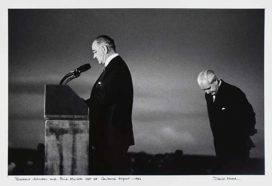 President Lyndon Johnson stands at a podium with microphones and Harold Holt stands behind him, head bowed. President Lyndon Johnson stands at a podium with microphones and Harold Holt stands behind him, head bowed.
