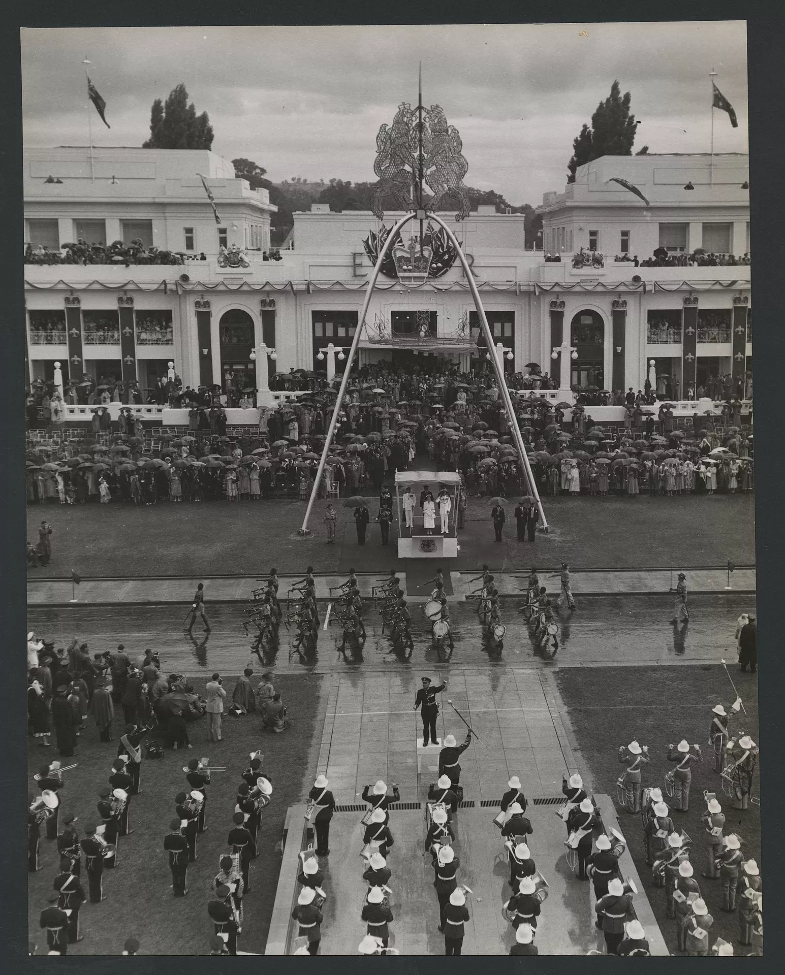Overhead view of Queen Elizabeth standing in a podium out the front of Old Parliament House, surrounded by a crowd holding umbrellas as troops march past. Overhead view of Queen Elizabeth standing in a podium out the front of Old Parliament House, surrounded by a crowd holding umbrellas as troops march past.
