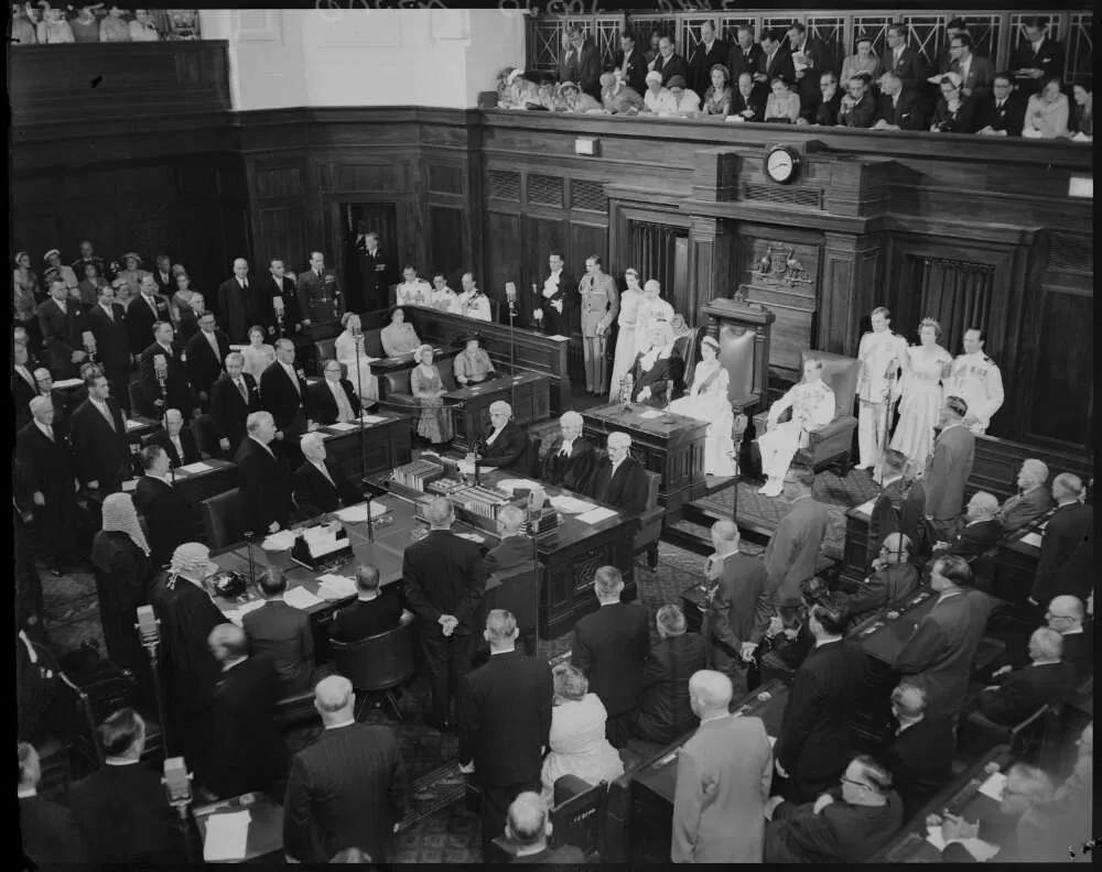 A black and white photo of the Senate Chamber filled with people standing up looking to an ornate chair at the front of the room where the Queen sits wearing a crown and a sash. A black and white photo of the Senate Chamber filled with people standing up looking to an ornate chair at the front of the room where the Queen sits wearing a crown and a sash.
