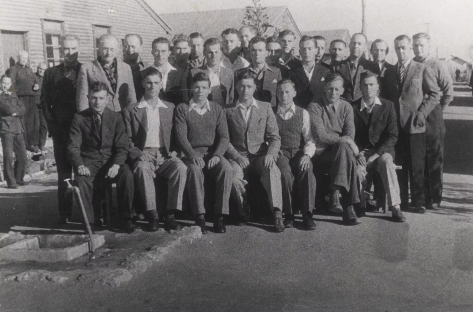 About 25 men in suits or smart casual clothing posing for a group photograph looking dignified towards the camera but not smiling. They are in a military camp in front of long wooden huts. About 25 men in suits or smart casual clothing posing for a group photograph looking dignified towards the camera but not smiling. They are in a military camp in front of long wooden huts.