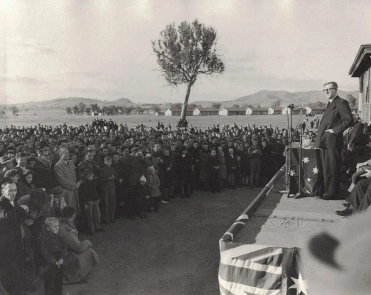 Arthur Calwell stands on a platform with microphones, addressing a crowd of people. In the distance is a row of long buildings. Arthur Calwell stands on a platform with microphones, addressing a crowd of people. In the distance is a row of long buildings.