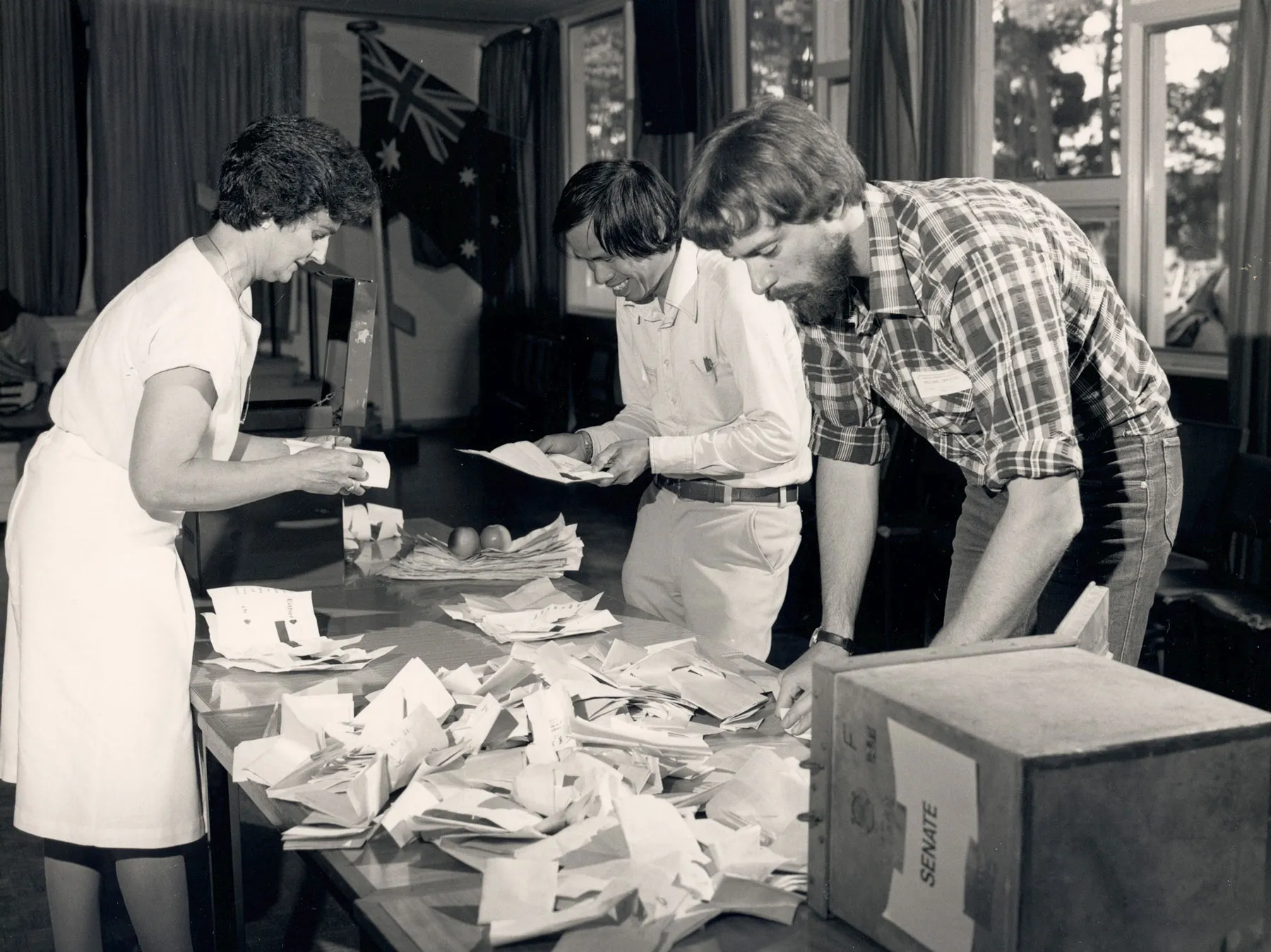 Three people stand around a table counting paper votes. There is a box titled 'Senate' on the table. Three people stand around a table counting paper votes. There is a box titled 'Senate' on the table.