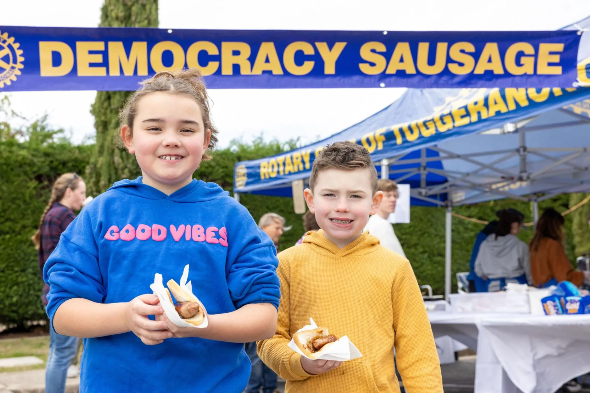 A girl wearing a blue hoodie with the text 'Good vibes' and a boy wearing a mustard hoodie each hold a sausage sandwich in front of an outdoor tent with a 'Democracy sausage' banner in the garden of Old Parliament House. A girl wearing a blue hoodie with the text 'Good vibes' and a boy wearing a mustard hoodie each hold a sausage sandwich in front of an outdoor tent with a 'Democracy sausage' banner in the garden of Old Parliament House.
