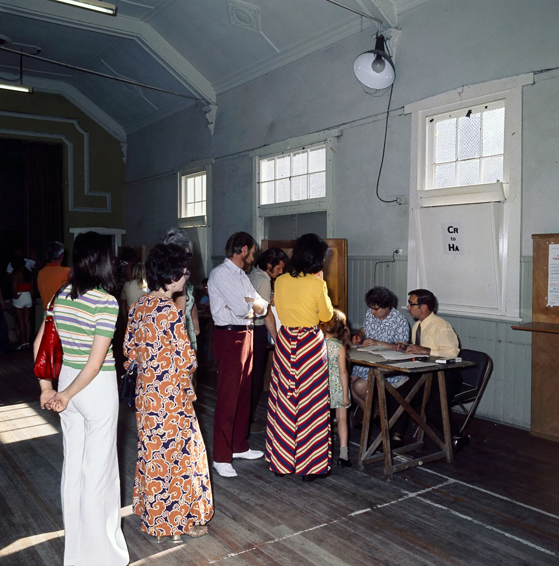 Men and women in colourful '70s clothes queue in a hall being used as a polling station. Election officials sit at wooden tables. Men and women in colourful '70s clothes queue in a hall being used as a polling station. Election officials sit at wooden tables.