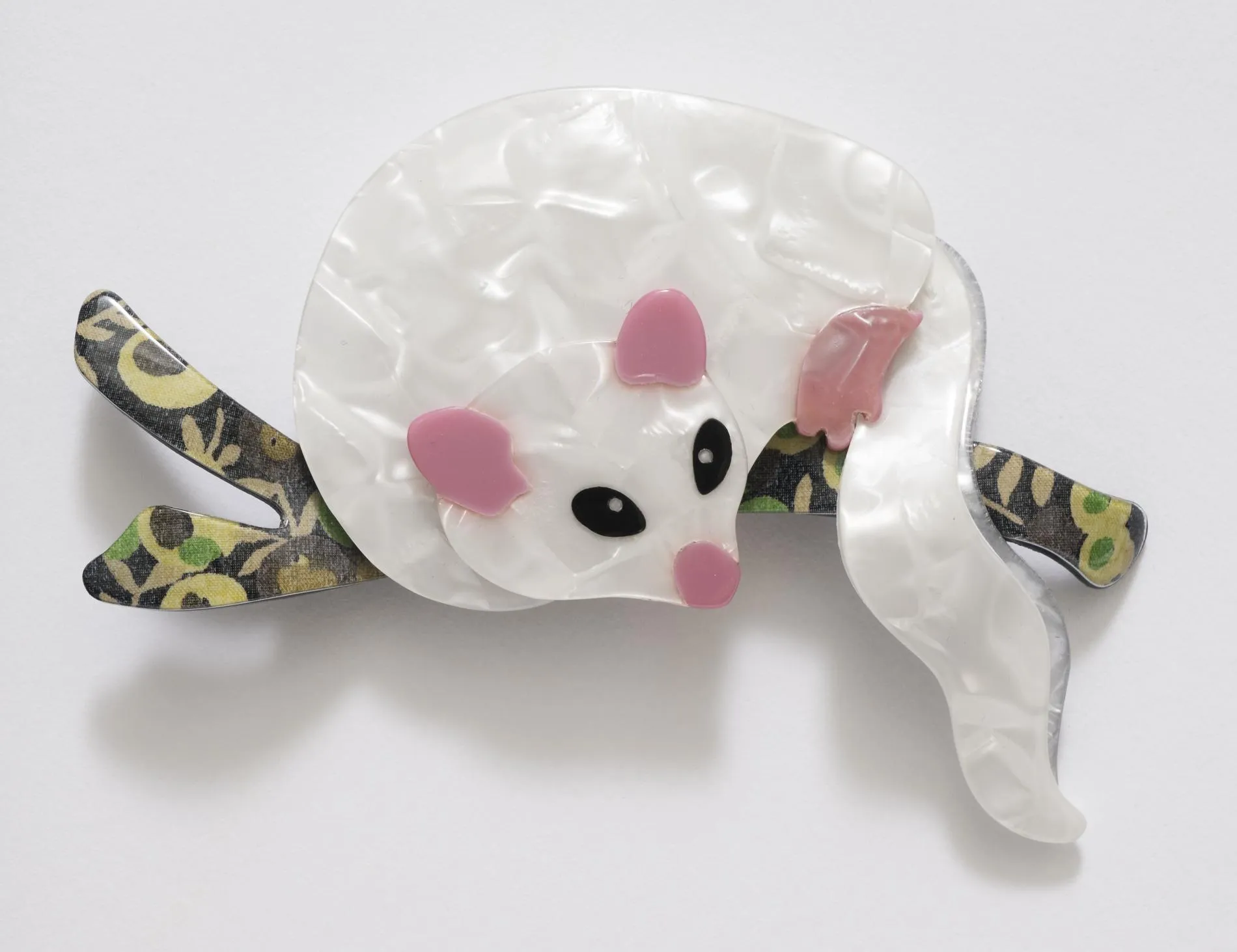 A celluloid possum brooch. The possum is white with pink ears, nose and feet, it sits on a black, green and gold branch. A celluloid possum brooch. The possum is white with pink ears, nose and feet, it sits on a black, green and gold branch.