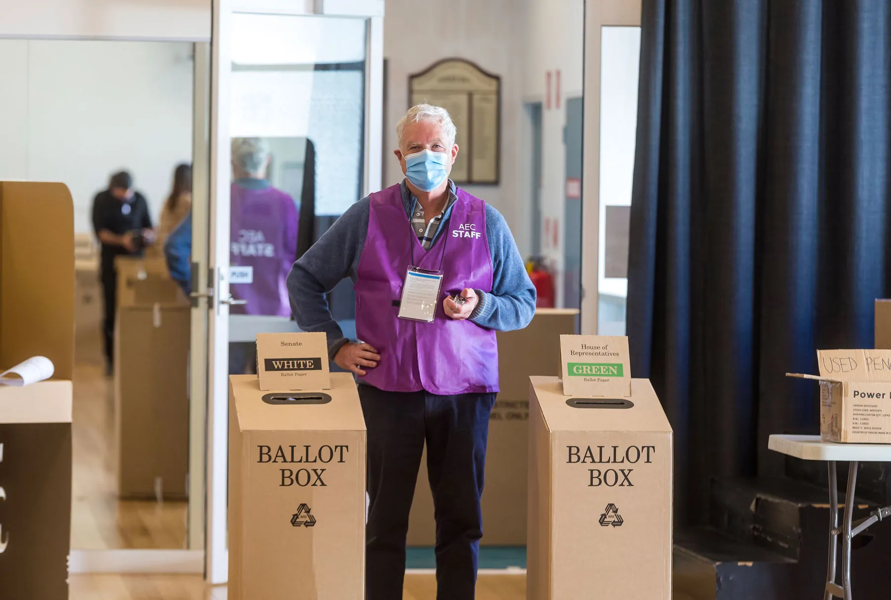 A man in a purple Australian Electoral Commission staff vest stands between two cardboard ballot boxes. A man in a purple Australian Electoral Commission staff vest stands between two cardboard ballot boxes.