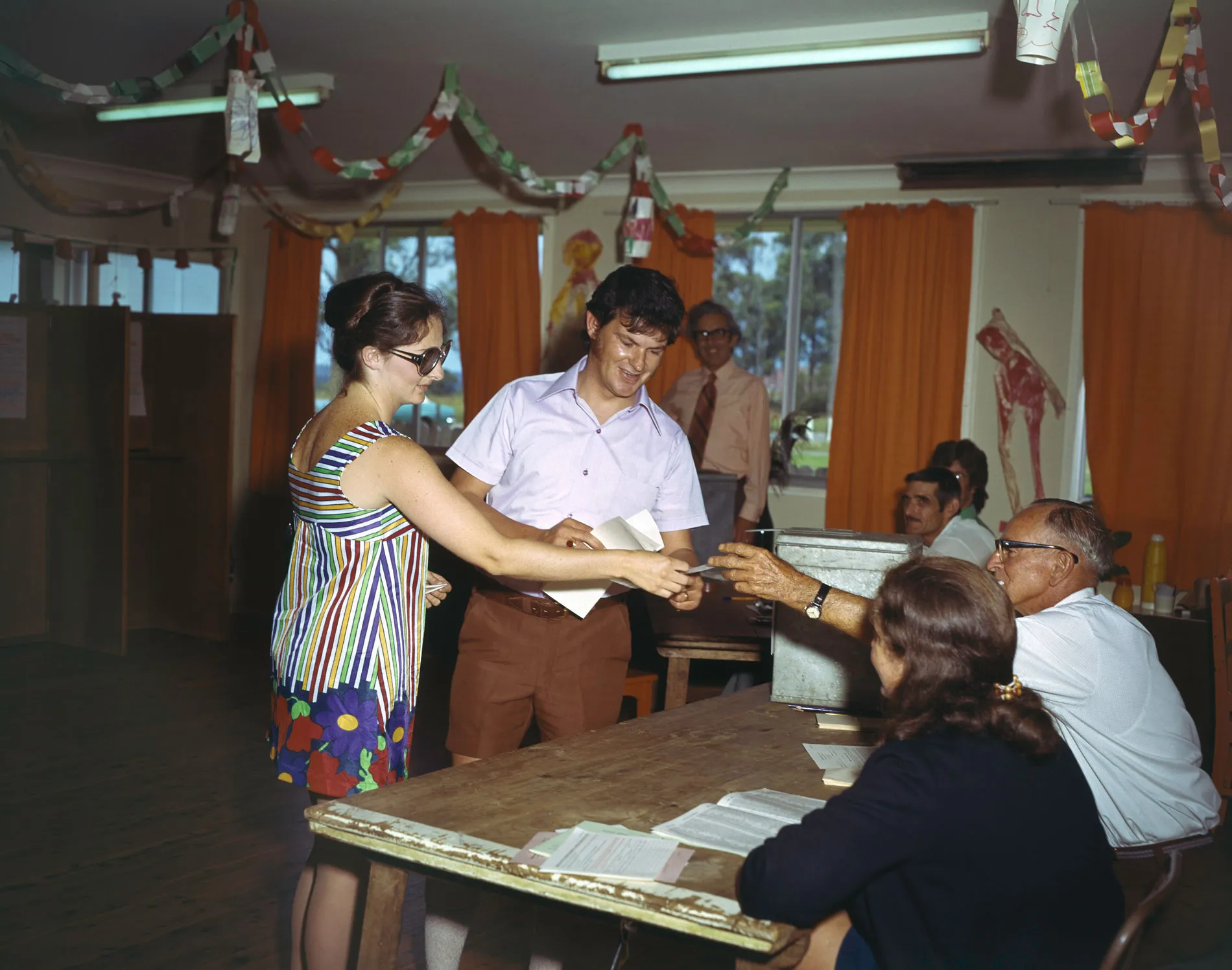 Two people being handed information at a polling booth in a hall with orange curtains.