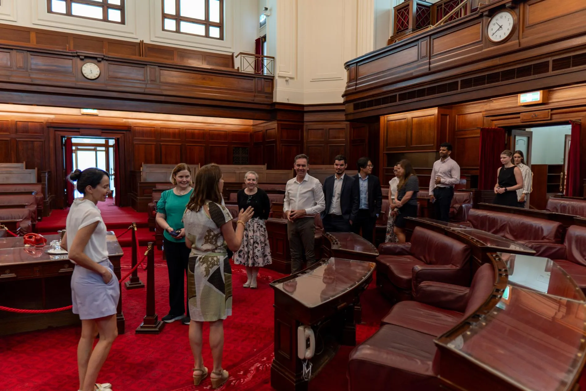 People on a tour in the Senate Chamber at Old Parliament House, it has red carpet and leather bench seats..