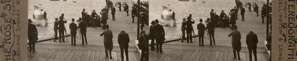 A black, white and grey historic photograph mounted on khaki grey card. An elevated view from above and behind small clusters of people who stand on a wooden pier. They are looking out at a boat that is headed towards the pier.  The label reads "The American Fleet in Australia. Rear-Admiral Sperry meeting the Prime Minister (Hon. Alfred Deakin) at St. Kilda." 1908.