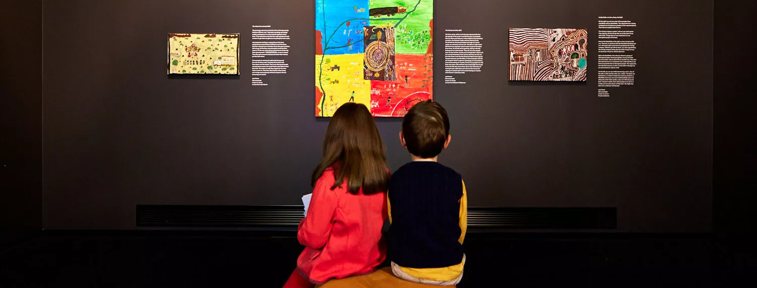 A boy and girl sit on a stool looking up at artworks from Jack Green. 