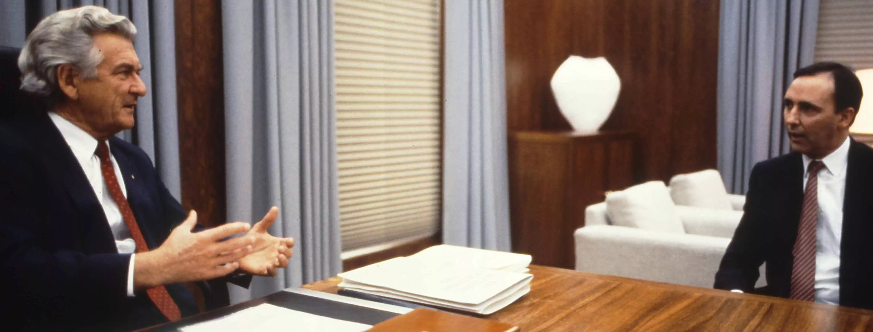 This colour photograph captures a conversation between Prime Minister Bob Hawke seated on the left and Treasurer Paul Keating seated on the right. The photo shows the corner of the desk with Hawke sitting behind the desk while Keating sits to the side with both men facing each other. Both are dressed in black suits with white shirts and red ties contrasting the pale blue curtains in the background.  