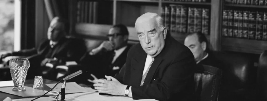 This black and white photograph shows Prime Minister Robert Menzies seated at a large table in the Government Party Room in 1965. He is conducting a press conference and there is a microphone, jug and water glass, and papers on the table. Behind him are three men seated in easy chairs and a bookcase filled with Hansards (volumes of parliamentary debates).  