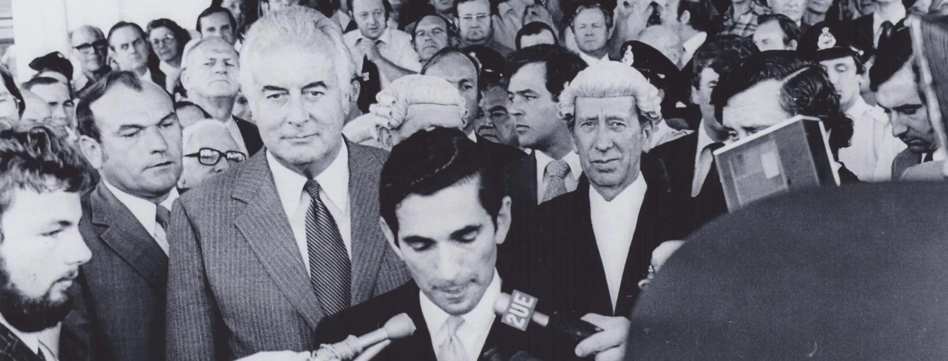 David Smith reads the dismissal proclamation outside Parliament House in 1975. Gough Whitlam stands behind him amongst a huge crowd. Reporters hold microphones.