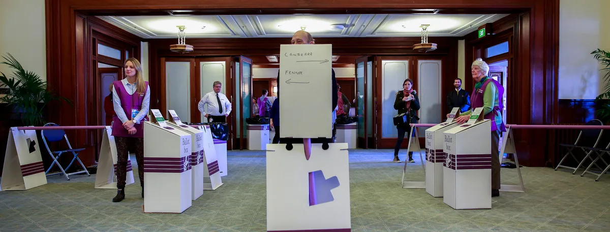 White ballot boxes for Canberra and Fenner are set up on green carpet in Old Parliament House. Australian Electoral Commission staff in purple vests stand next to the boxes. White ballot boxes for Canberra and Fenner are set up on green carpet in Old Parliament House. Australian Electoral Commission staff in purple vests stand next to the boxes.