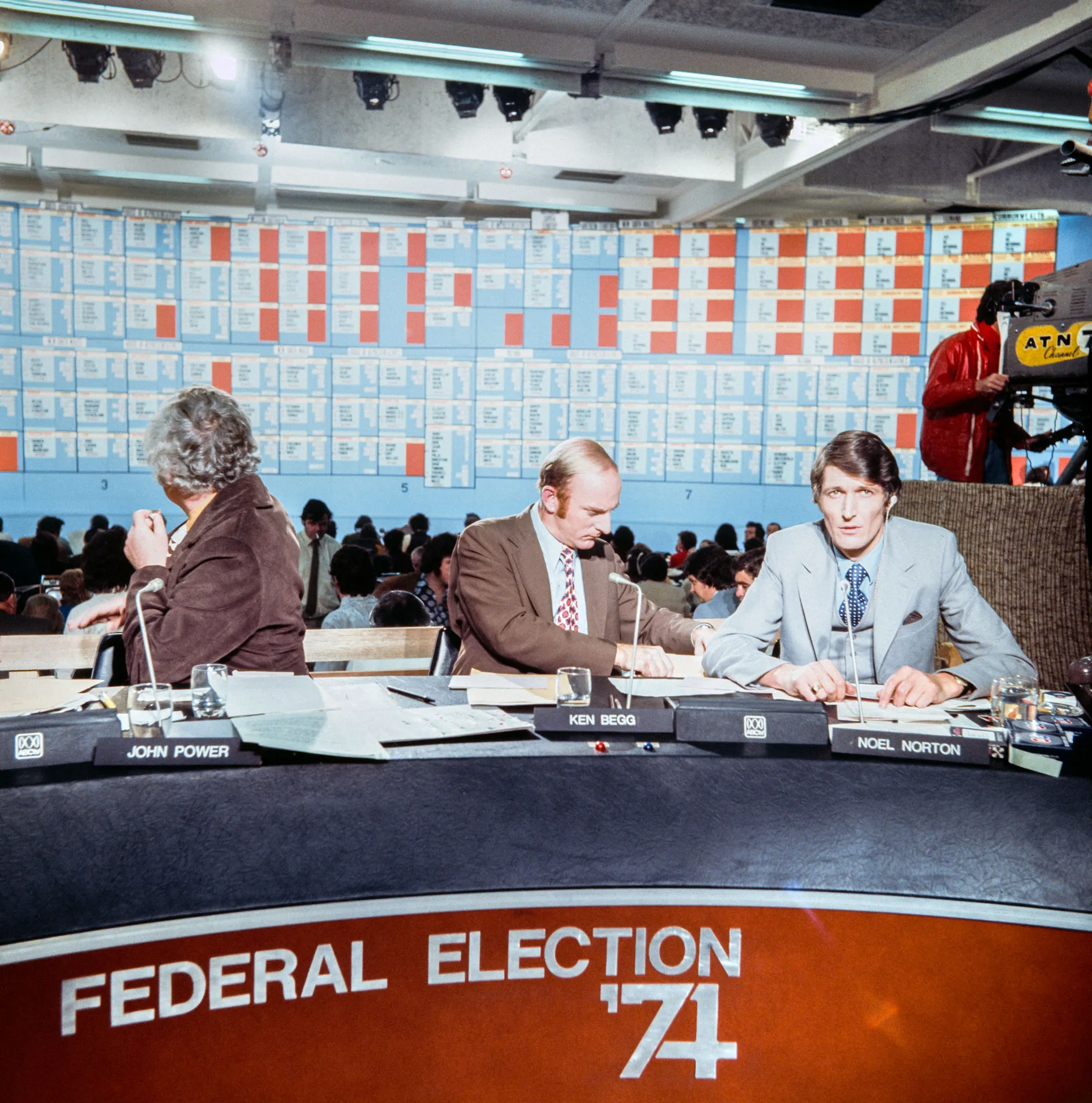 Three reporters sit at a Federal election '74 desk in front of a giant tally board. Three reporters sit at a Federal election '74 desk in front of a giant tally board.