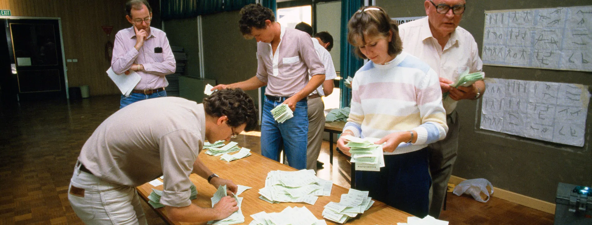 Men and women count and sort green ballot papers on a wooden table in the 1984 election.