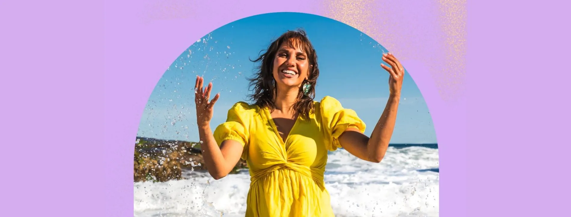 Kirli Saunders wears a yellow dress and stands in the waves on a beach.