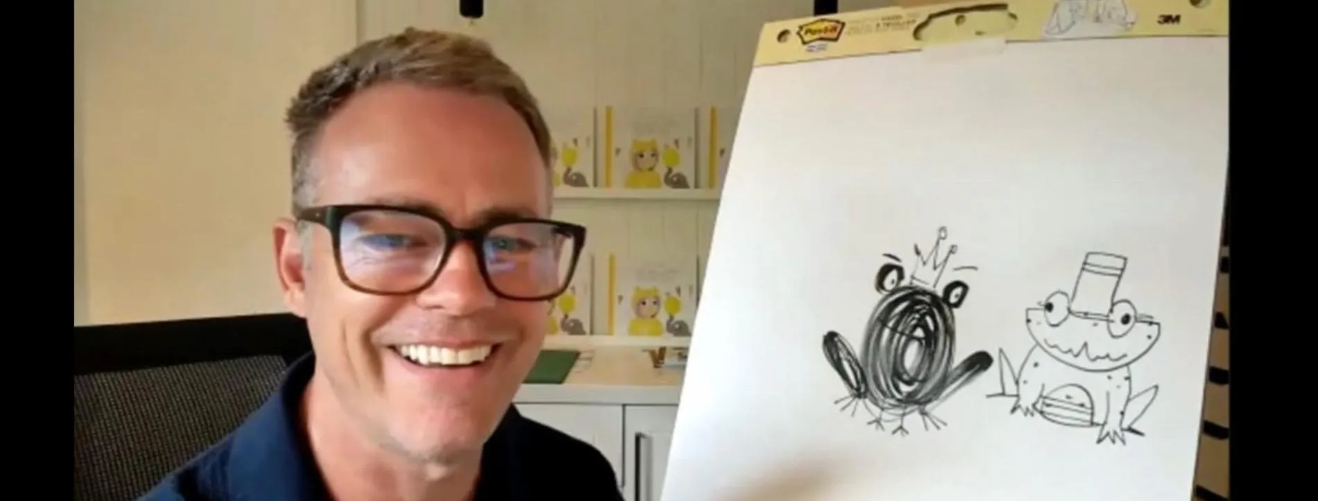 A man wearing glasses is smiling at the camera. There is a drawing next to him on an easel.