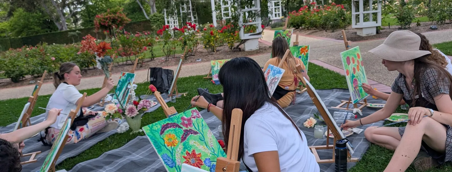 People sit on the grass in the rose gardens at Old Parliament House painting floral artworks.
