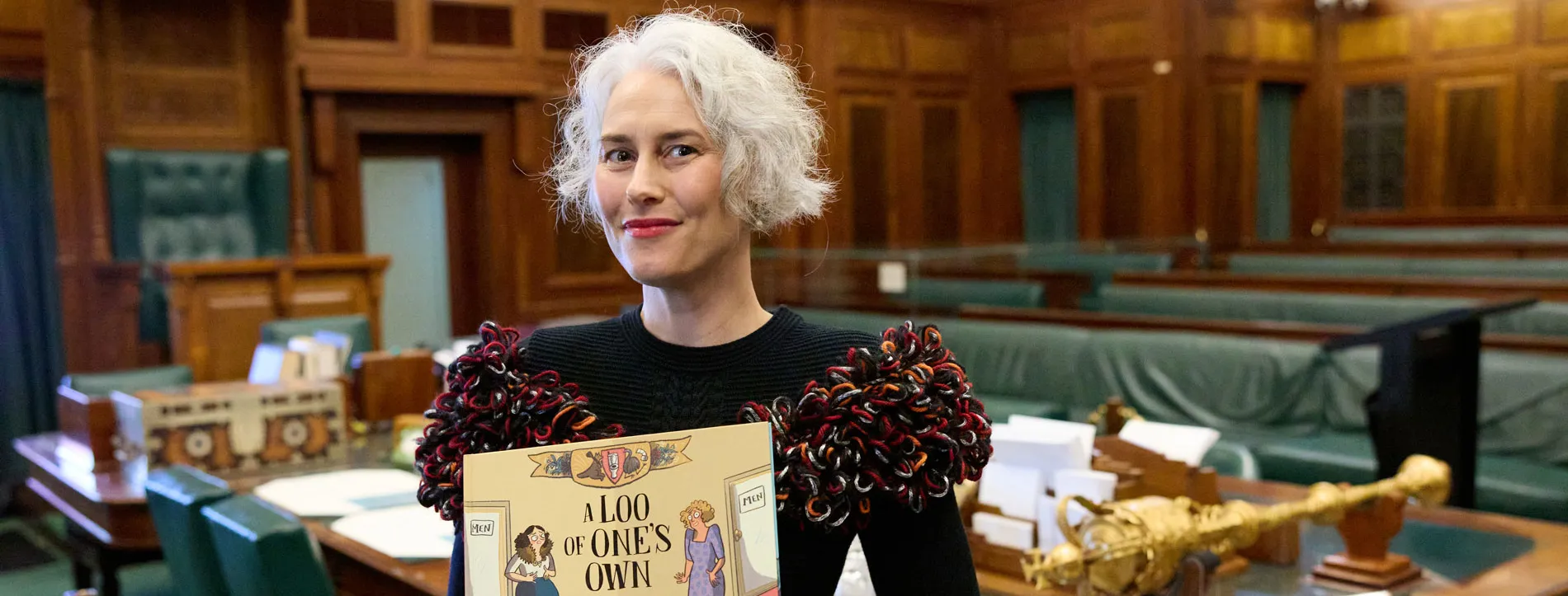 Author Eleri Harris holds a book standing in the green Senate Chamber of Old Parliament House.