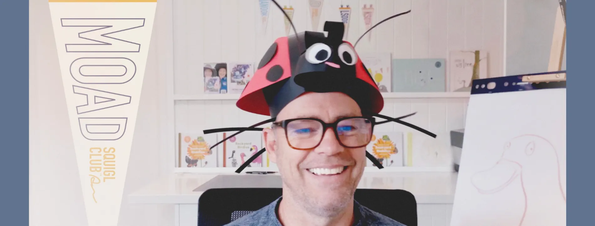 Andy Geppert in front some bookshelves with his books on them, with a lady bug made of paper on his head.