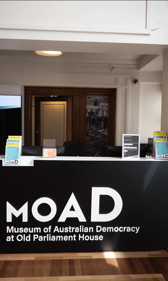 A front desk with pamphlets on top of it and a large black sign with the words 'MoAD Museum of Australian Democracy at Old Parliament House.' A front desk with pamphlets on top of it and a large black sign with the words 'MoAD Museum of Australian Democracy at Old Parliament House.'