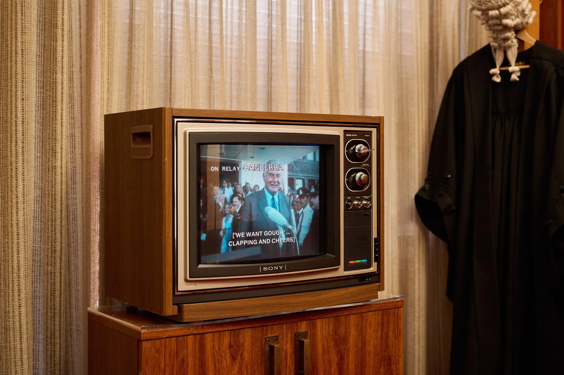 A wood-framed TV with an image of Gough Whitlam sits on a wood cabinet. A black gown and white wig hang next to the TV. A wood-framed TV with an image of Gough Whitlam sits on a wood cabinet. A black gown and white wig hang next to the TV.