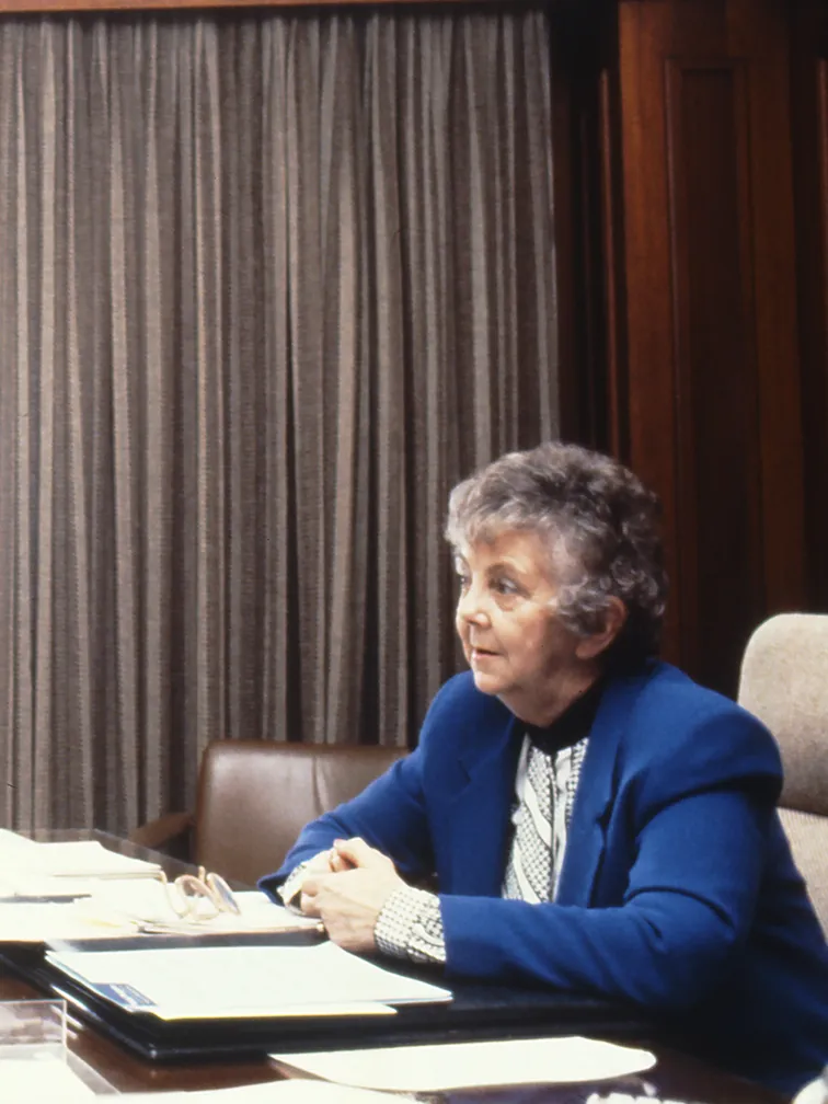 This professional colour photograph shows Principal Private Secretary John Porter and Speaker Joan Child sitting across from each other at the Speaker’s desk. John is wearing a dark suit, white shirt and dark tie and Joan is in a white blouse and vibrant blue jacket. They are discussing the daily program for the House of Representatives Chamber. There is a vase of roses, stationery and paperwork on the desk. In the background the curtains have been drawn to reveal a large east-facing window.