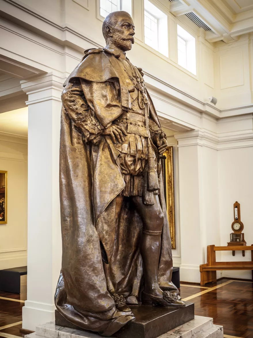 Generations of journalists have leant against the toe of King George V, polishing it to a high shine. A bronze statue of a man with a cloak on a marble pillar in a room with timber floors.
