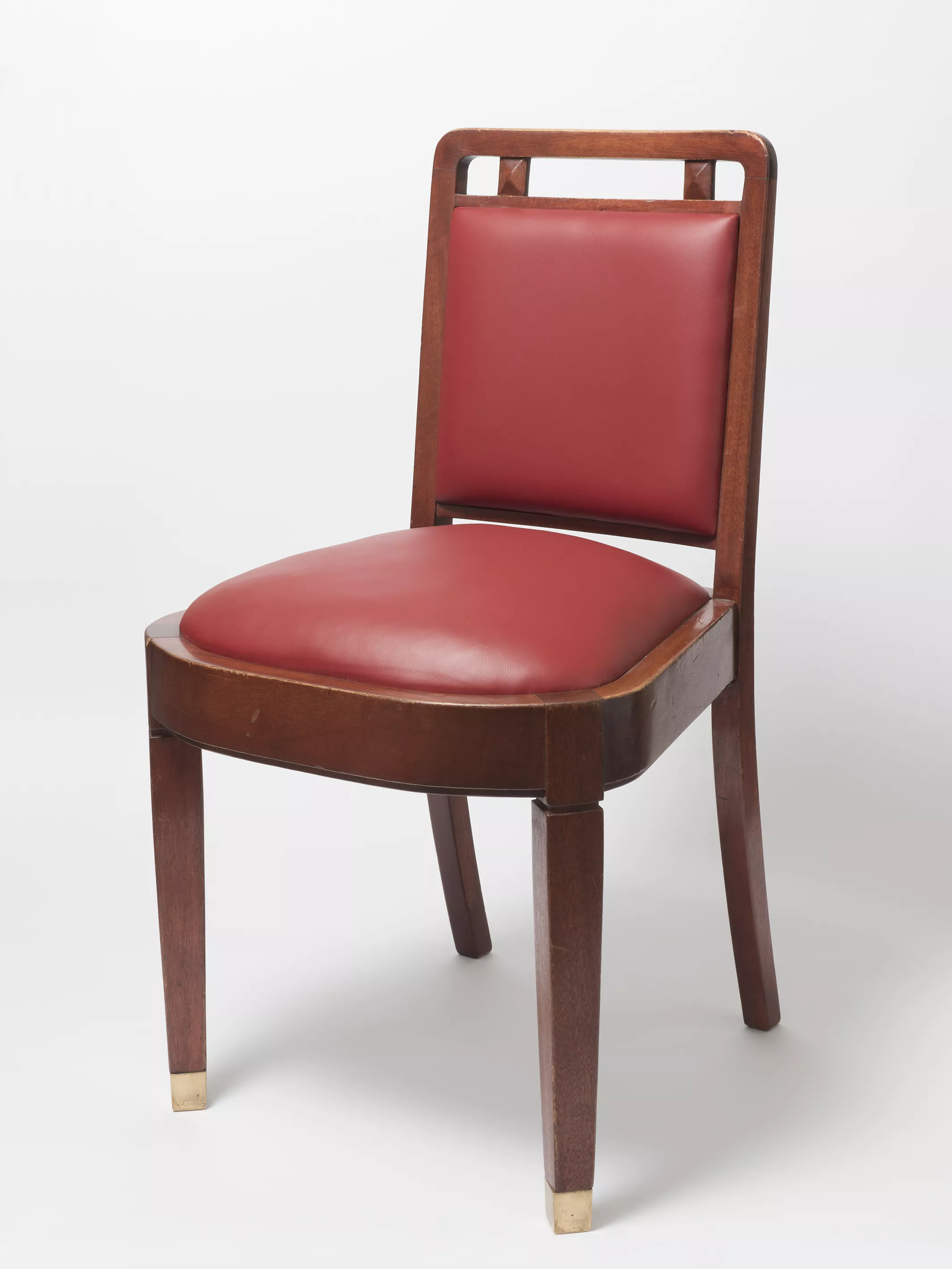 A wooden chair with cherry red leather seat cushion and back, and square legs with protective brass caps on the base of each leg. A wooden chair with cherry red leather seat cushion and back, and square legs with protective brass caps on the base of each leg.