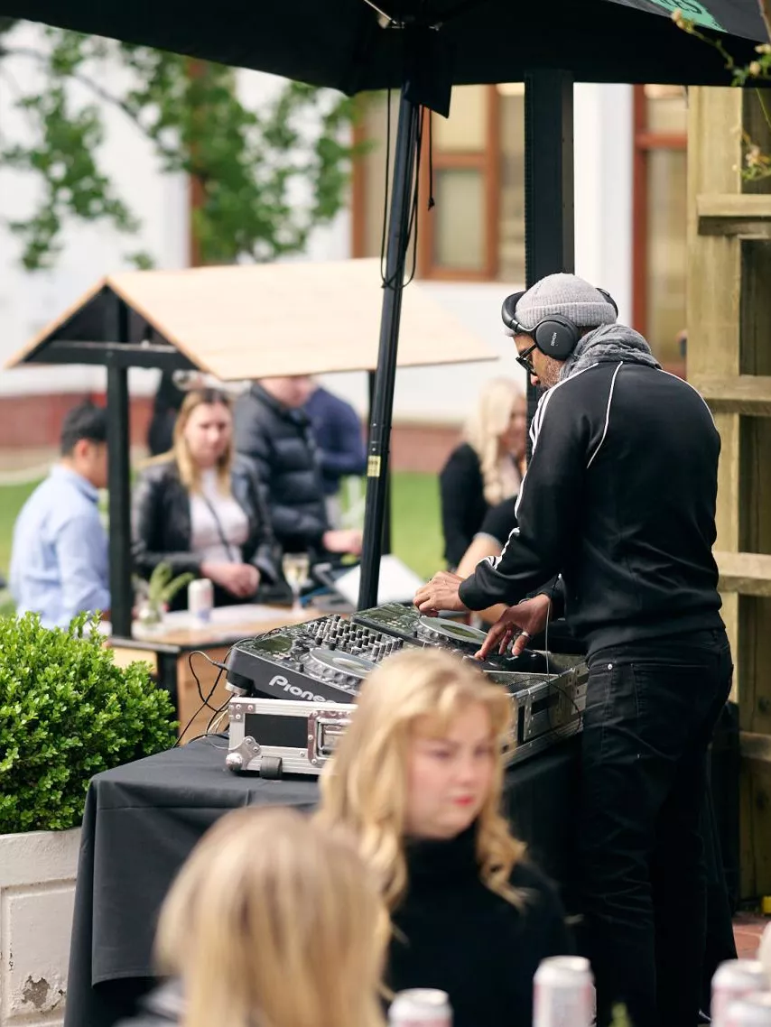 A man wearing headphones operating DJ equipment in the House of Representatives courtyard, surrounded by groups of people socialising and drinking cans of beer.