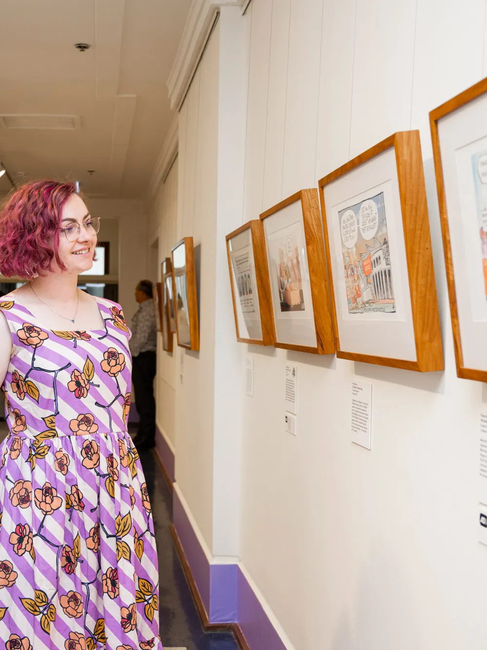 Two smiling people look at framed cartoons on the wall of an exhibition. 