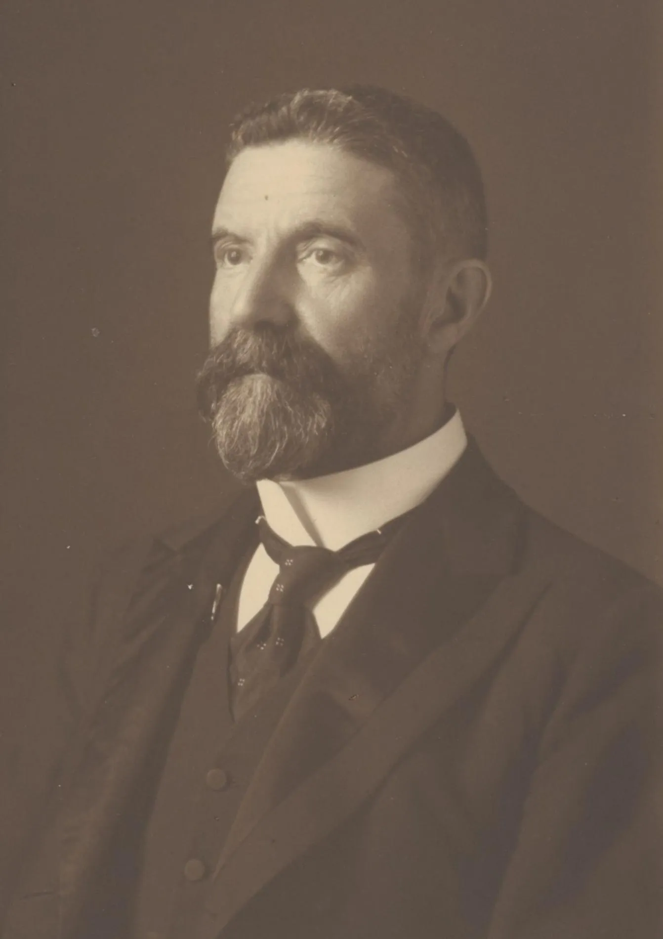 A black and white headshot of Alfred Deakin. He's looking slightly left, in a suit and tie. He's got a short haircut with a well kept beard and moustache.