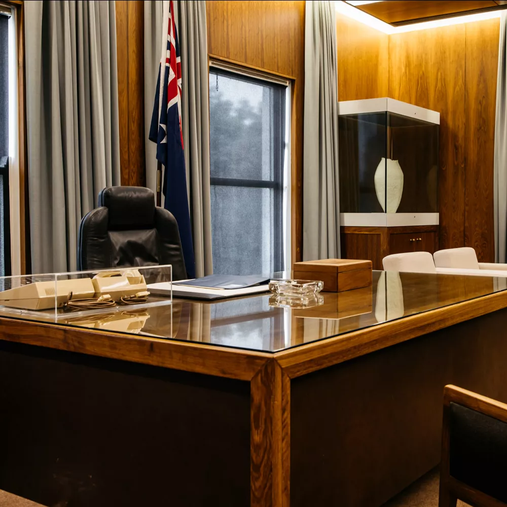 A black bean wooden desk with a glass cover in a room with an Australian flag, leather chair and grey curtins. A black bean wooden desk with a glass cover in a room with an Australian flag, leather chair and grey curtins.