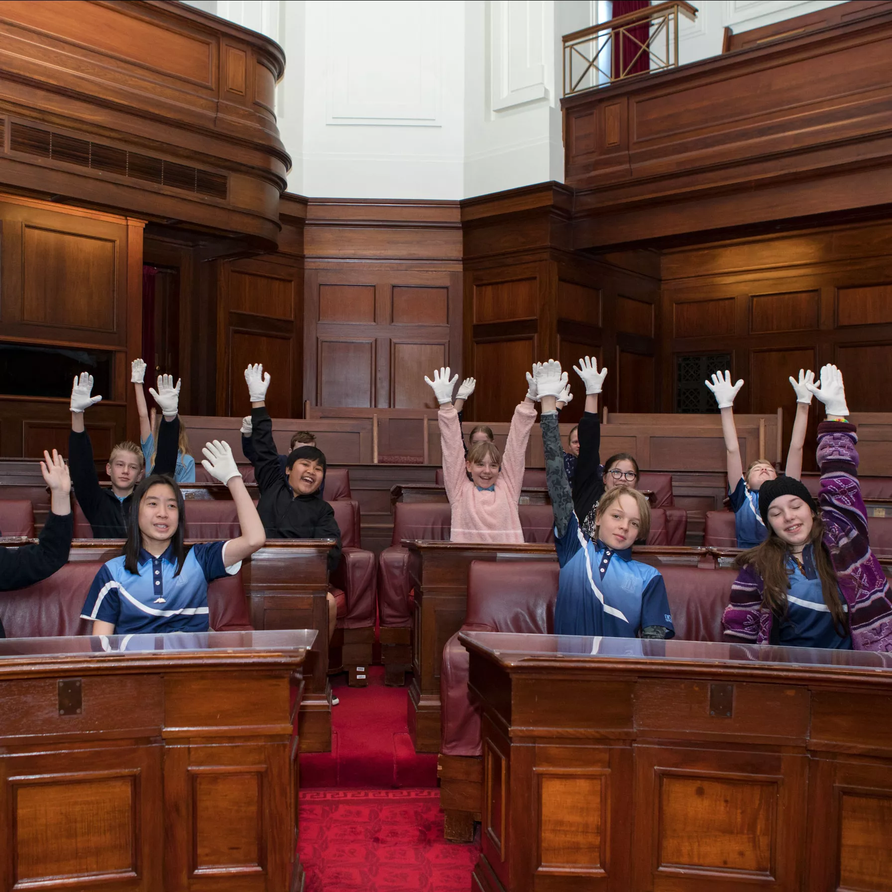 School children raise their hands while sitting in the Senate Chamber which has red carpet and wood panelled benches in a U-shape. 