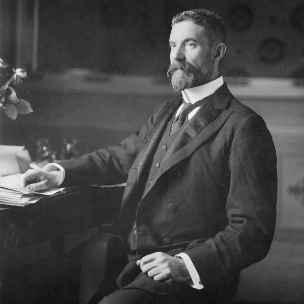 Alfred Deakin, wearing a three-piece suit and seated at a desk covered in paper documents.