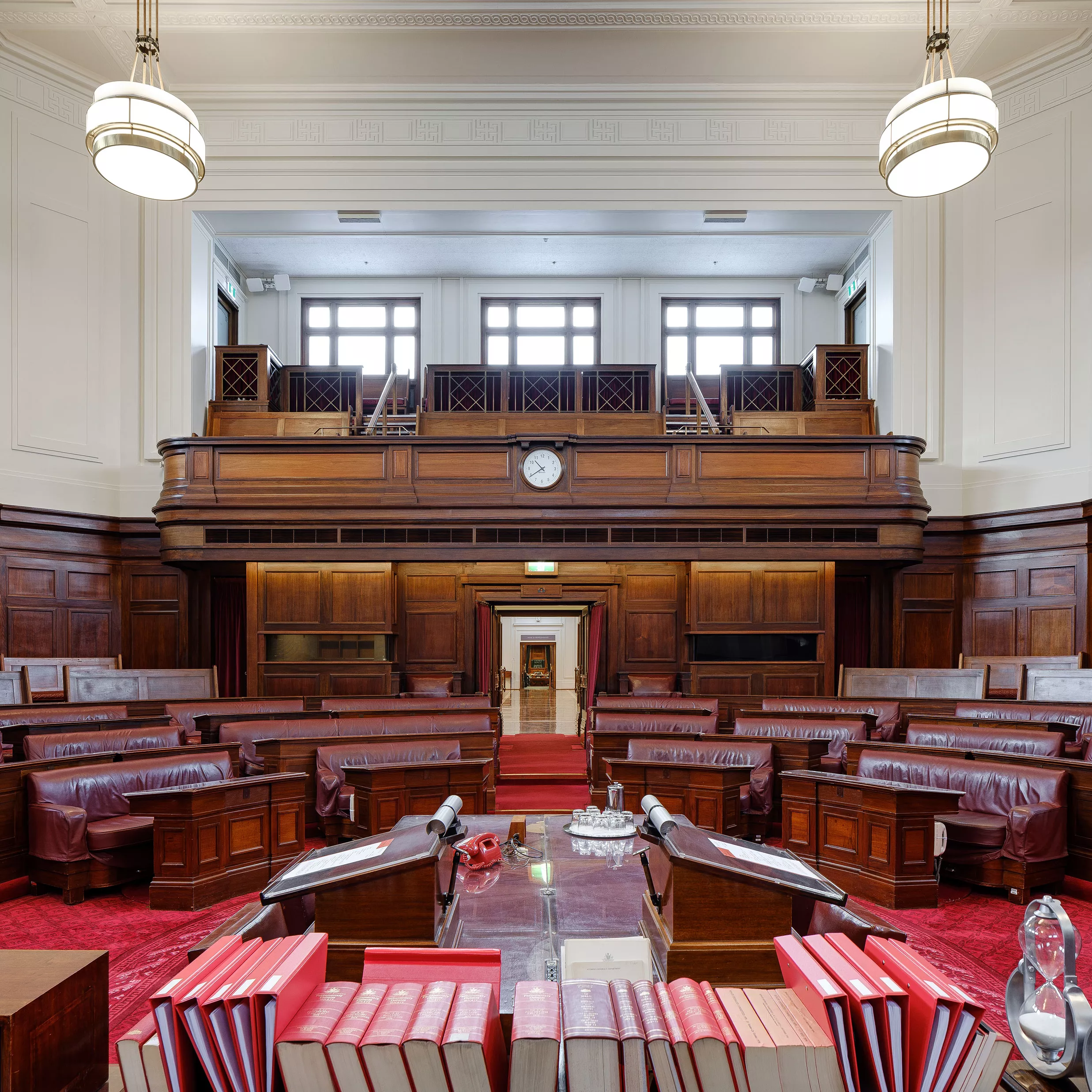 A view out to the Senate chamber. In the foreground are red leather bound books on a long table. Red carpet and red leather chairs in a horseshoe shape with timber panels and art deco lights. 