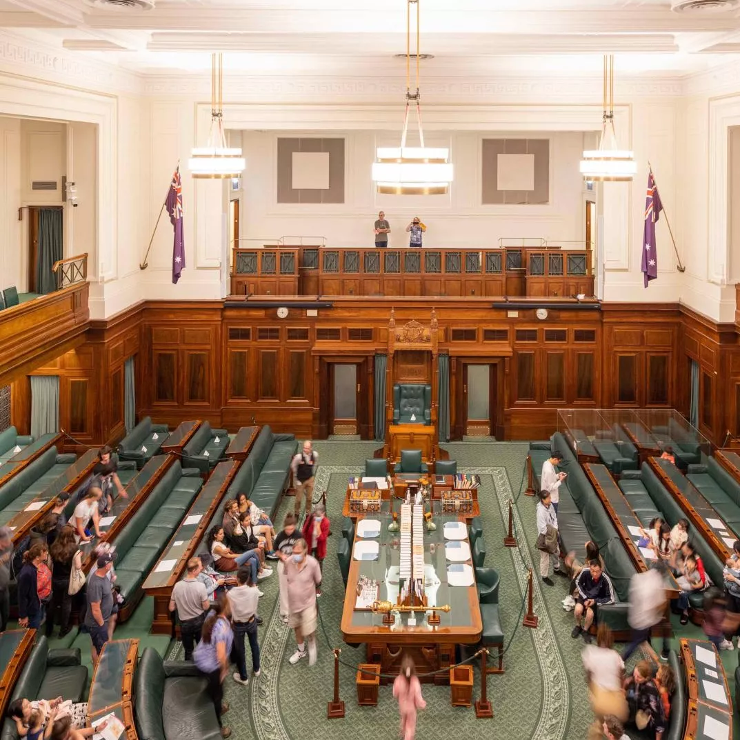 A view looking down onto the House of Representatives Chamber which has green carpet and green leather and timber benches in a U-shape surrounding a central table. 