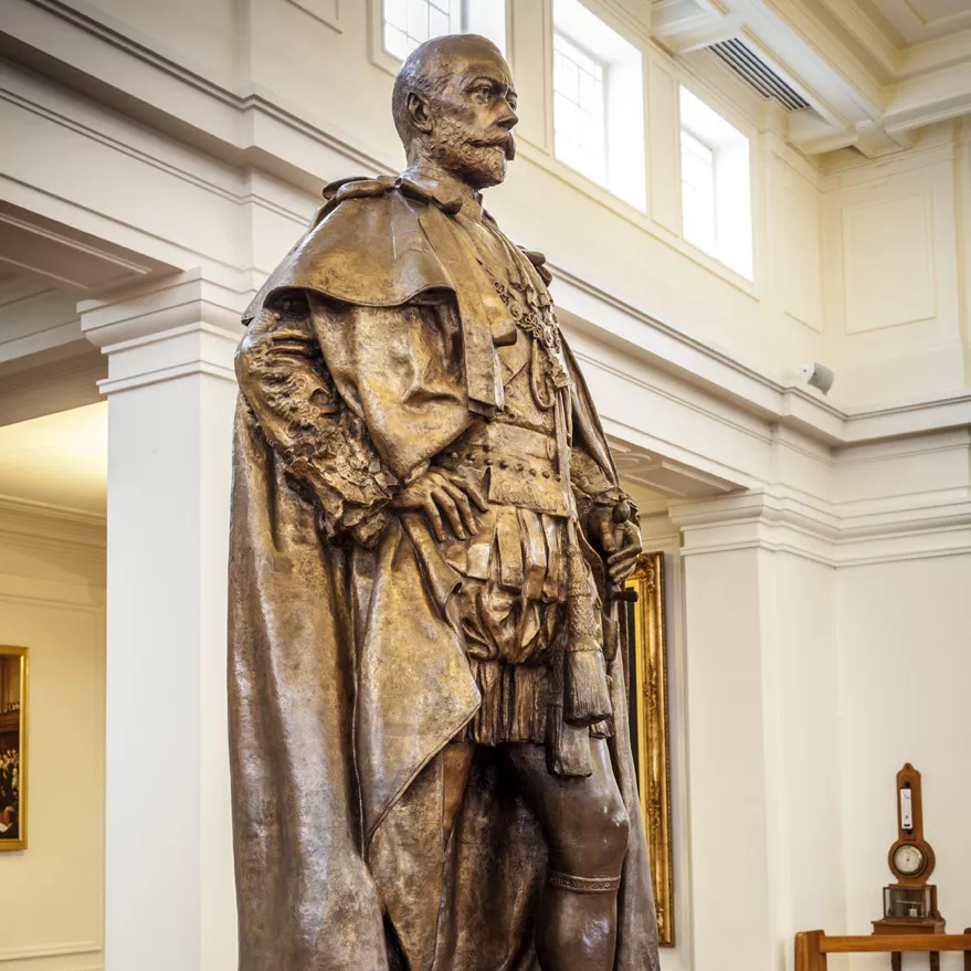 Generations of journalists have leant against the toe of King George V, polishing it to a high shine. A bronze statue of a man with a cloak on a marble pillar in a room with timber floors.