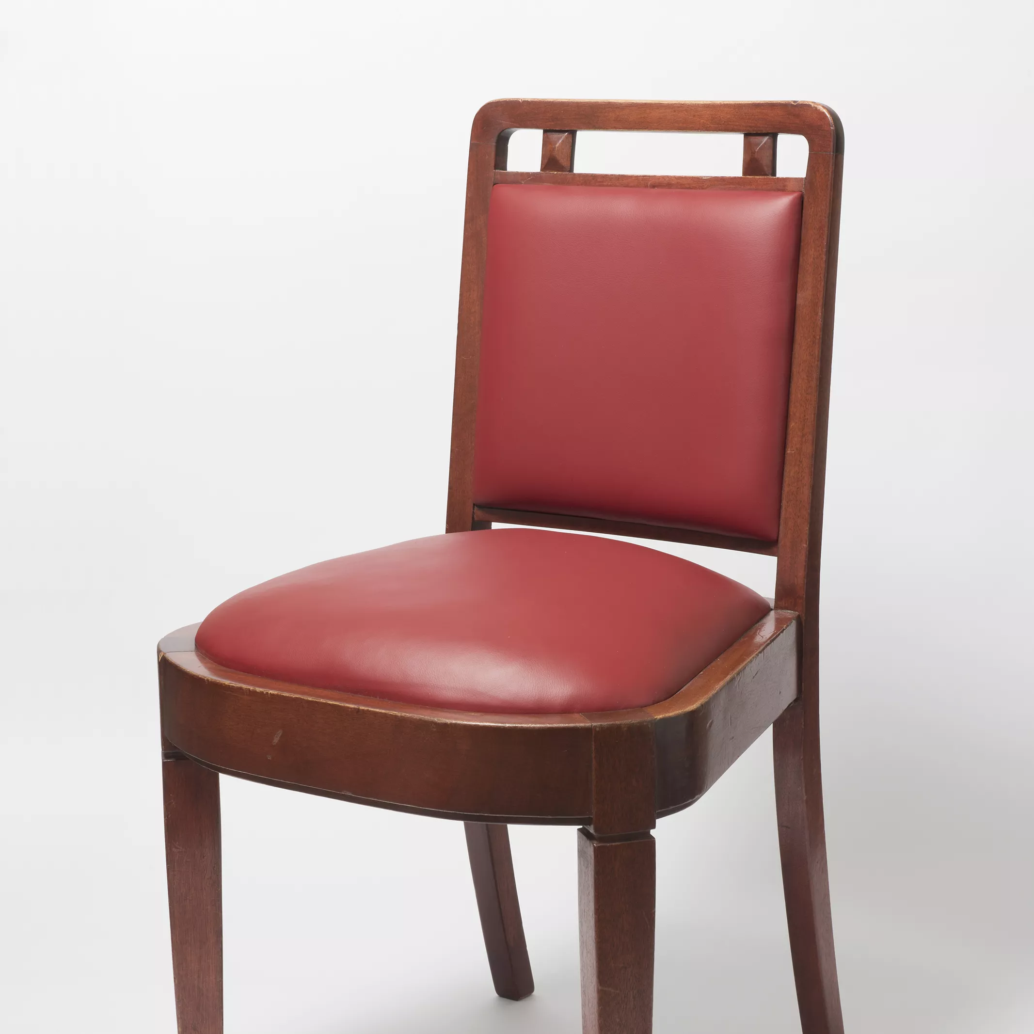 A wooden chair with cherry red leather seat cushion and back, and square legs with protective brass caps on the base of each leg. 