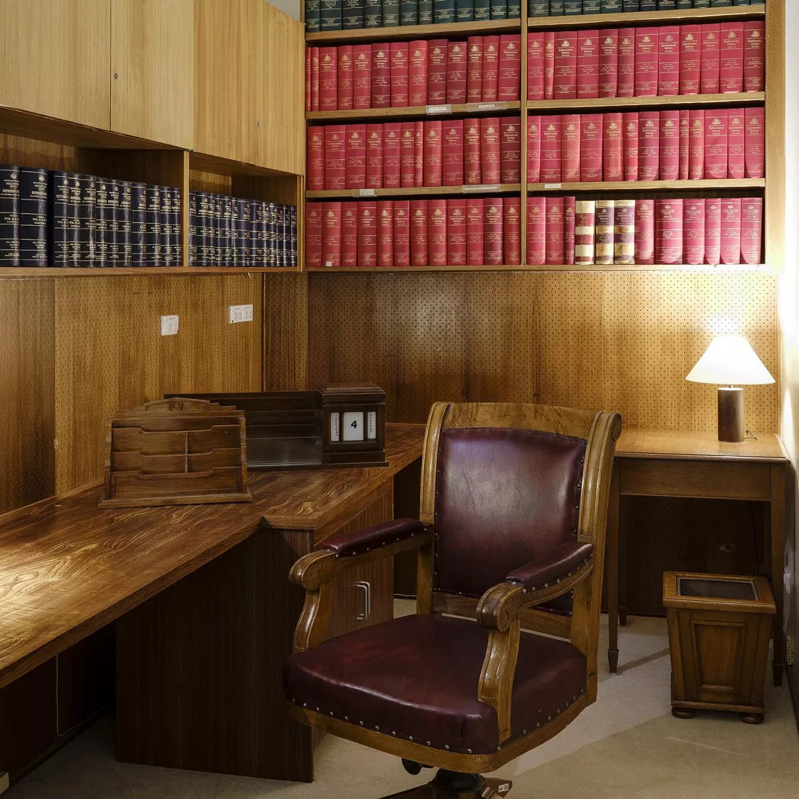 A leather-cushioned wooden swivel chair, next to a long wooden desk with a lamp and desk calendar; on two walls there are inbuilt bookcases with rows of bound books. A leather-cushioned wooden swivel chair, next to a long wooden desk with a lamp and desk calendar; on two walls there are inbuilt bookcases with rows of bound books.