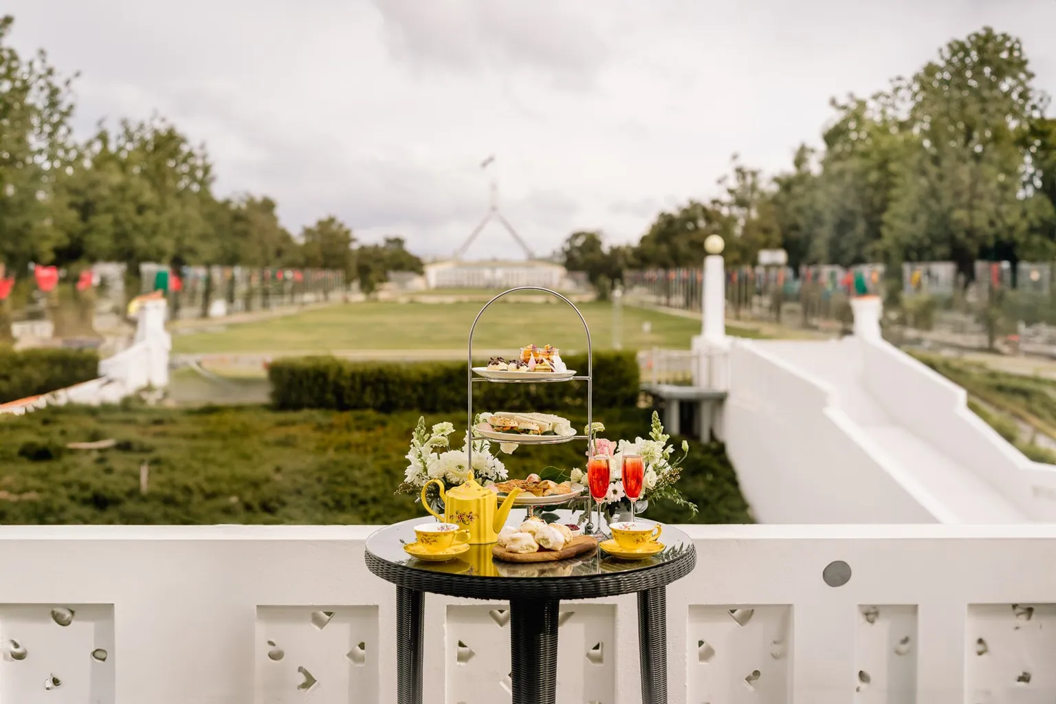 A table is set with a high tea tower filled with cakes and sandwiches is on the balcony of Old Parliament House. Australian Parliament House is in the background along with green lawns.