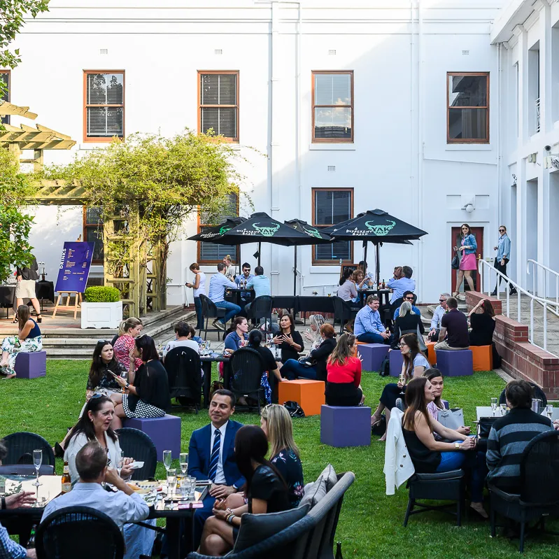 People sit at tables and on stools in the Old Parliament House courtyard having drinks.