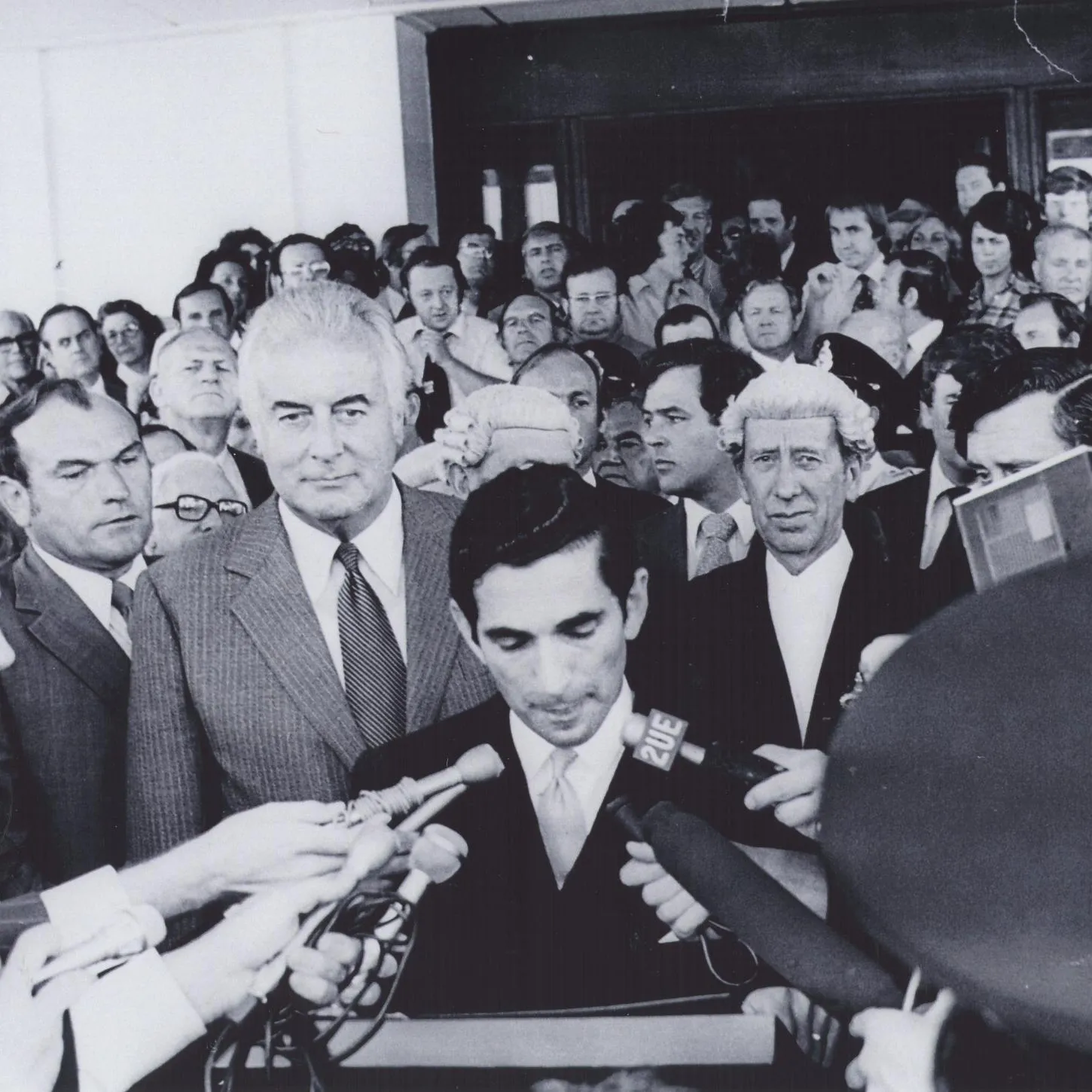 David Smith reads the dismissal proclamation outside Parliament House in 1975. Gough Whitlam stands behind him amongst a huge crowd. Reporters hold microphones.