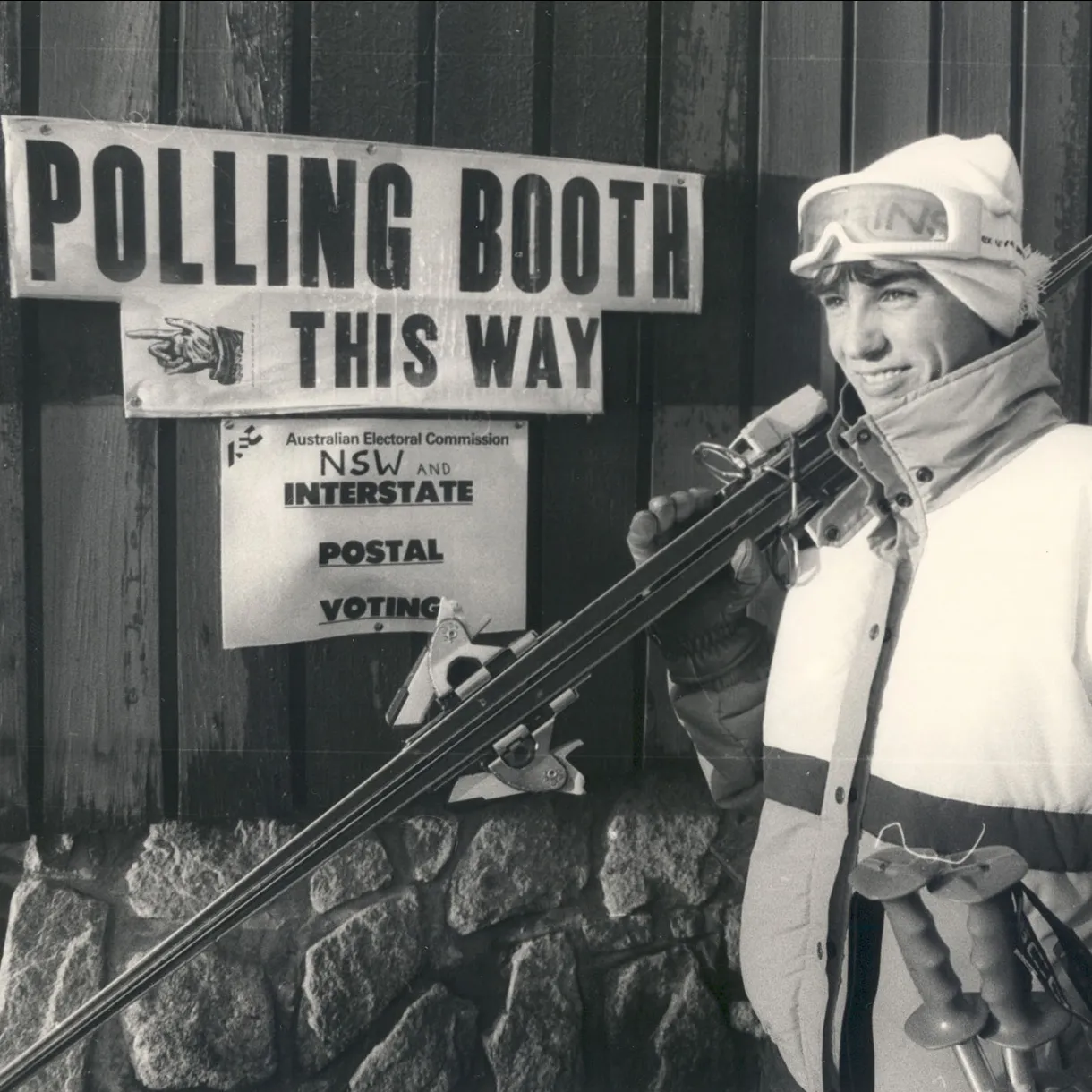 A man wearing vintage ski clothes and carrying skis stands outside a building with a 'Polling booth this way' sign. A man wearing vintage ski clothes and carrying skis stands outside a building with a 'Polling booth this way' sign.