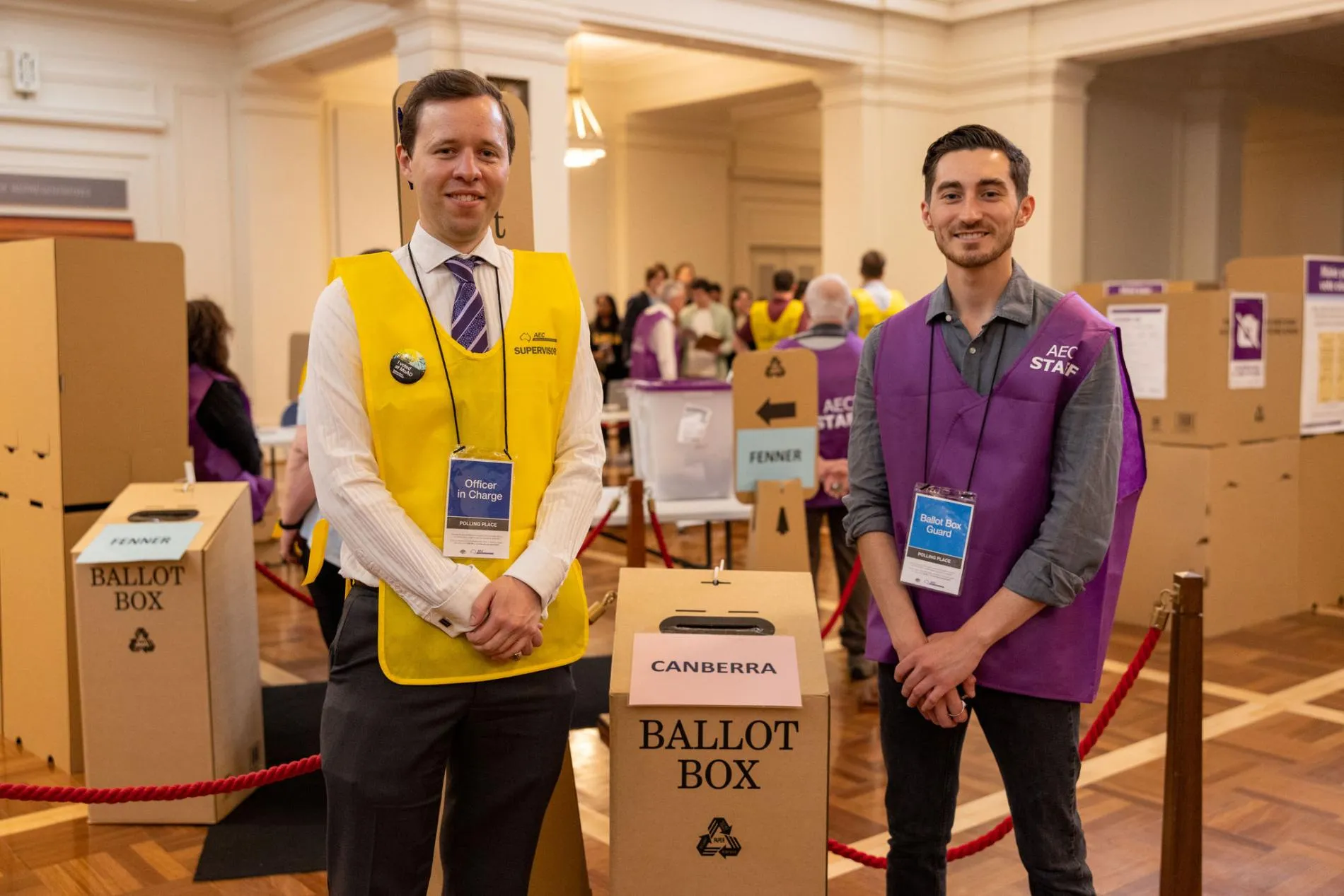 Two AEC officials, one in a yellow vest and one in a purple vest, stand beside a cardboard ballot box for Canberra in King's Hall in Old Parliament House. Two AEC officials, one in a yellow vest and one in a purple vest, stand beside a cardboard ballot box for Canberra in King's Hall in Old Parliament House.