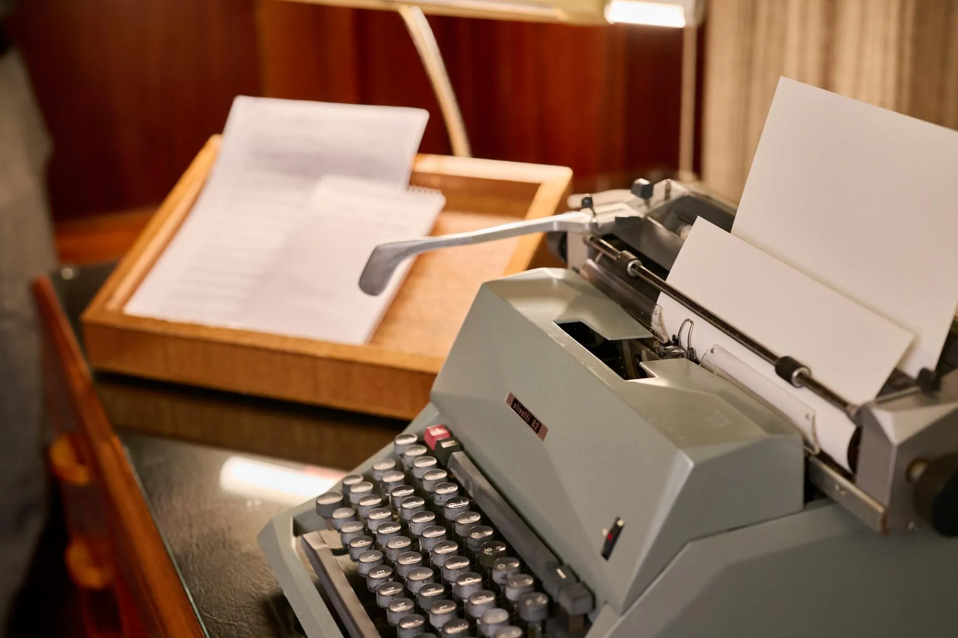 An old-fashioned grey typewriter and wooden document tray with paper in it sits on green leather-topped desk.