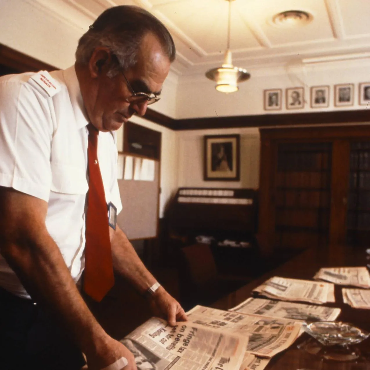 An attendant arranges the daily newspapers in the Senate Opposition Party Room, 1988.
Photograph by Robert McFarlane, Department of the House of Representatives A man wearing a white shirt and red tie arranges newspapers on a table in Old Parliament House.